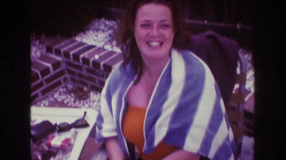 The adult woman smiles widely toward the camera in this 1975 Boston 8mm frame, seated outdoors with a striped towel, orange swimsuit, brick wall, and patio items nearby.