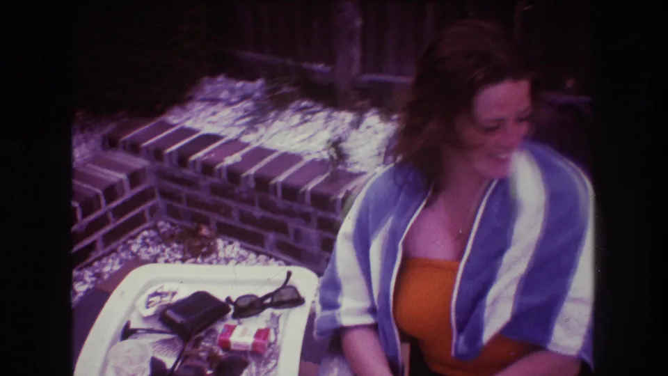 The 1975 Boston home-movie frame shows the woman looking down and laughing, wrapped in a striped towel beside a white tray holding sunglasses and a small dark wallet or case.