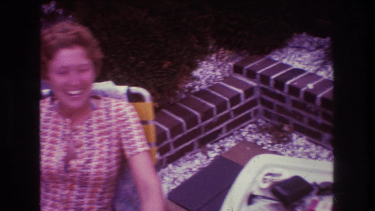 A woman with short red hair smiles while seated outdoors on a patio in Boston home-movie footage from 1975.
