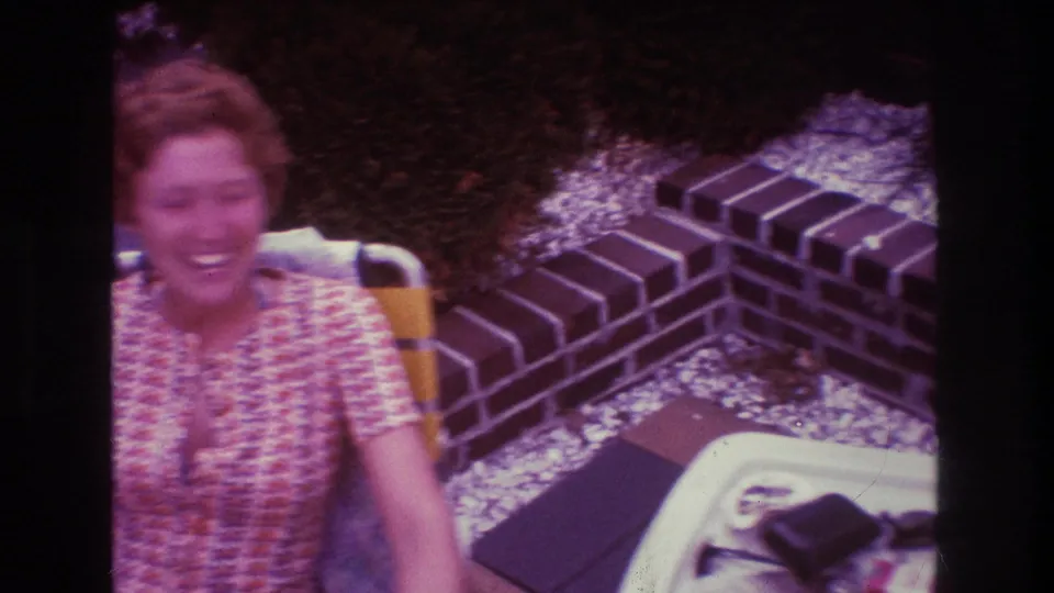 A woman with short red hair smiles while seated outdoors on a patio in Boston home-movie footage from 1975.
