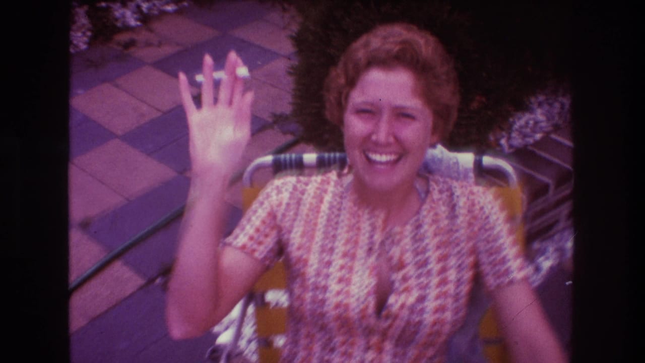Woman in a patterned dress waves toward the camera from a patio chair.