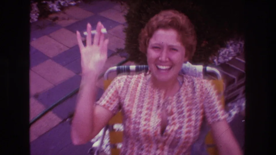 The seated woman waves up toward the camera while smiling on a checkered patio in 1975 Boston home-movie footage.
