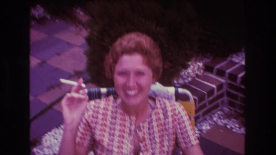 A seated woman smiles and holds a cigarette near her shoulder during an outdoor gathering in Boston, 1975.