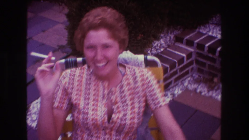 The woman continues smiling with a cigarette in hand, framed by brick edging, patio paving, and greenery in the 1975 home-movie scene.