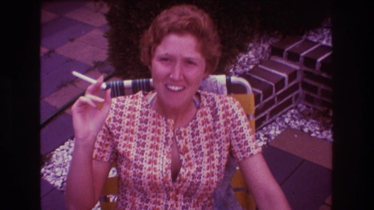 Adult woman in a patterned dress sits outdoors in a yellow lawn chair in 1975 Boston home-movie footage, holding a cigarette beside patio paving and a brick wall.