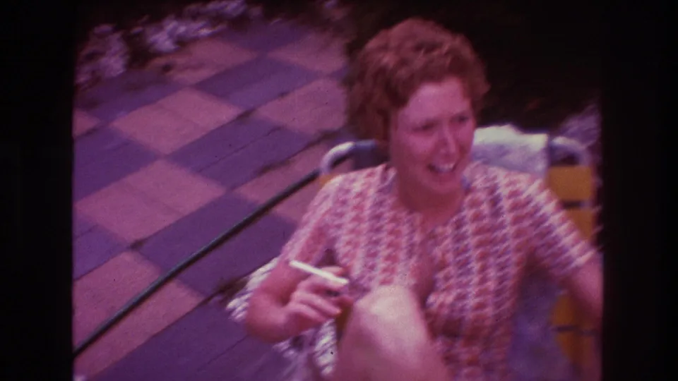 Adult woman laughs in a yellow lawn chair, holding a cigarette as the handheld 8mm camera catches motion blur over the patio setting.
