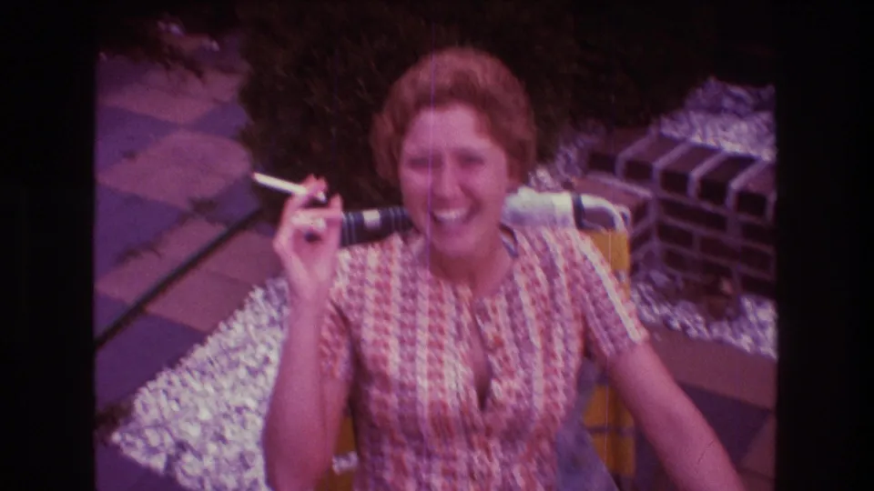 Adult woman in 1975 Boston home-movie footage laughs toward the camera while raising a cigarette, seated outdoors near patio pavers, white gravel, shrubs, and brickwork.