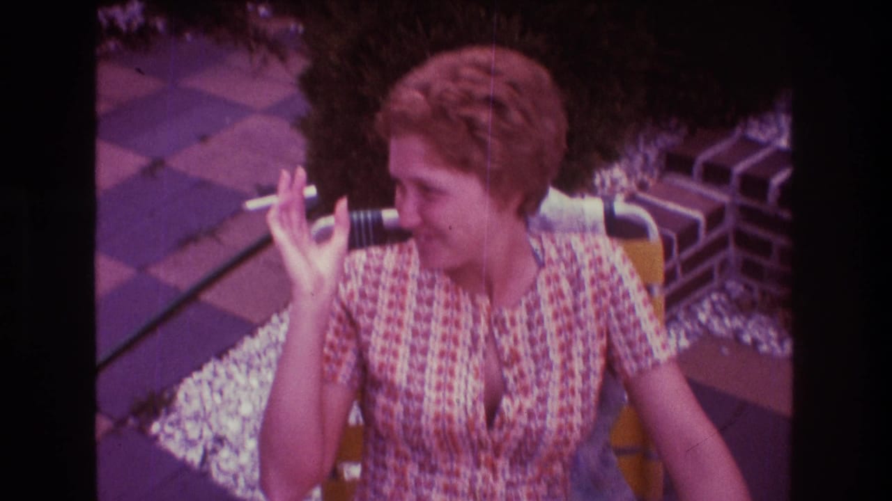 In supported 1975 Boston home-movie footage, a woman in a patterned dress sits outdoors holding a cigarette beside brick edging and patio paving.