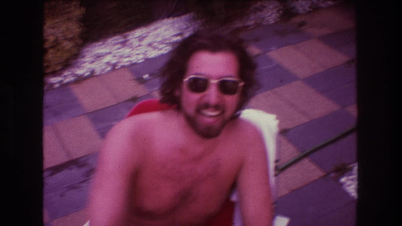 Shirtless man wearing sunglasses laughs while seated outdoors.