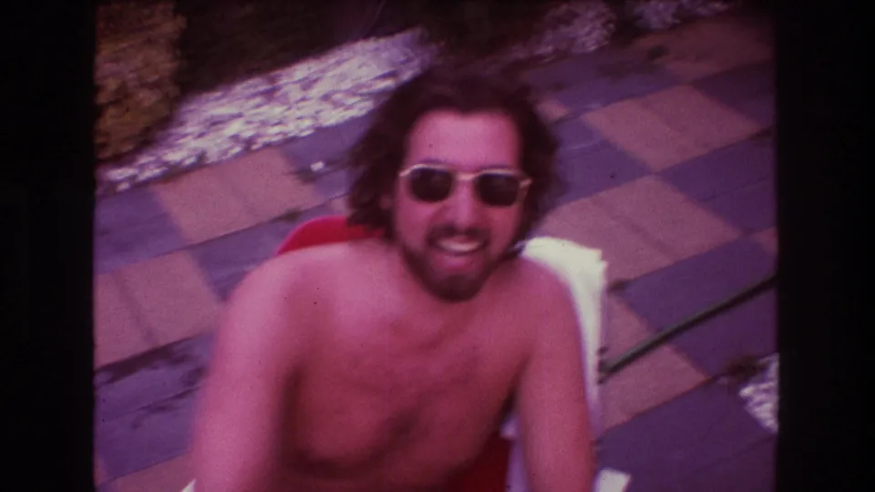 A shirtless adult man with long hair and sunglasses looks up toward the handheld camera while seated on a red patio chair in the 1975 Boston home-movie scene.