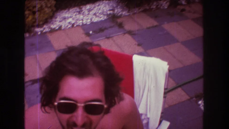 The camera moves close to the shirtless man in sunglasses as he leans forward from a red chair, with patio paving and a white towel visible around him.