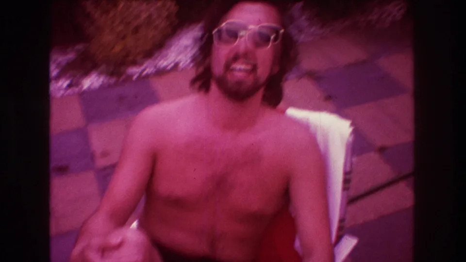 The shirtless man with sunglasses grins broadly and gestures upward toward the camera while seated outdoors on the checkered patio.