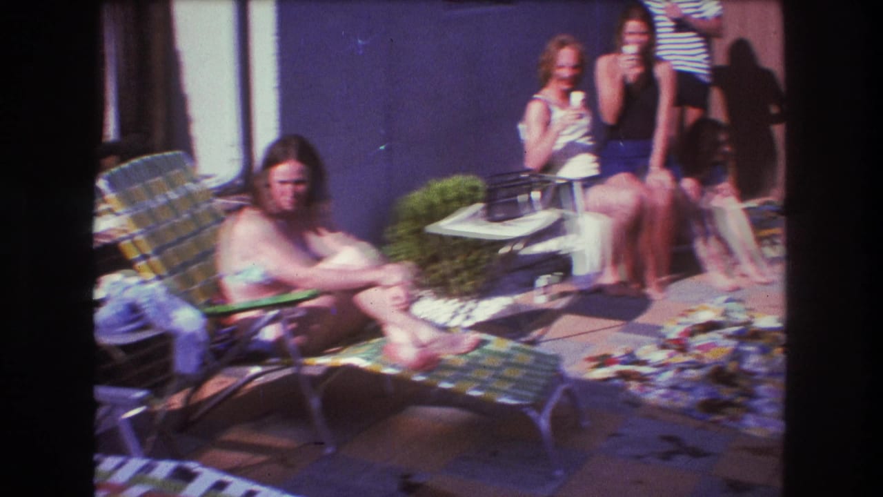 1975 Boston home-movie frame showing young adults in swimsuits and summer clothes relaxing in a backyard beside lawn chairs, blankets, a purple wall, and a wooden fence.