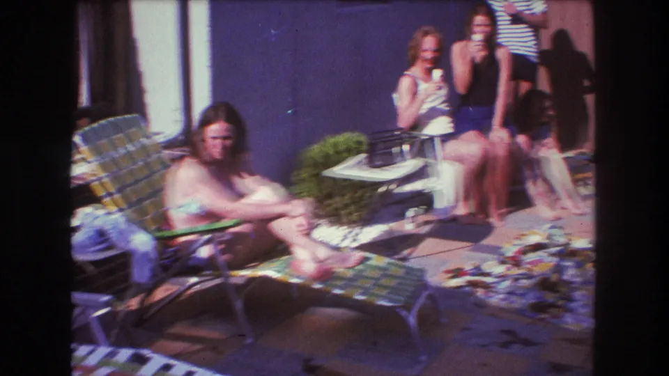 1975 Boston home-movie frame showing young adults in swimsuits and summer clothes relaxing in a backyard beside lawn chairs, blankets, a purple wall, and a wooden fence.