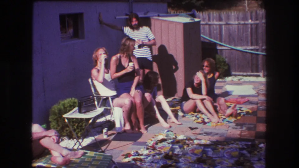 1975 Boston home-movie frame showing friends in casual summer clothing and swimsuits seated around backyard chairs and blankets, with a man standing behind them.