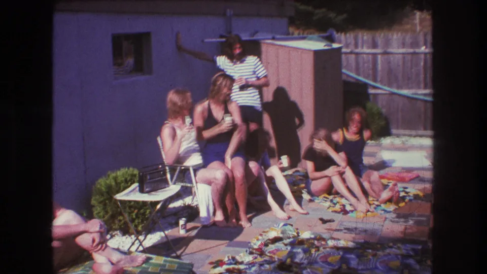 1975 Boston home-movie frame of young adults seated and standing in a sunny backyard patio area, with visible folding chairs, colorful blankets, a purple wall, a shed, and a wooden fence.