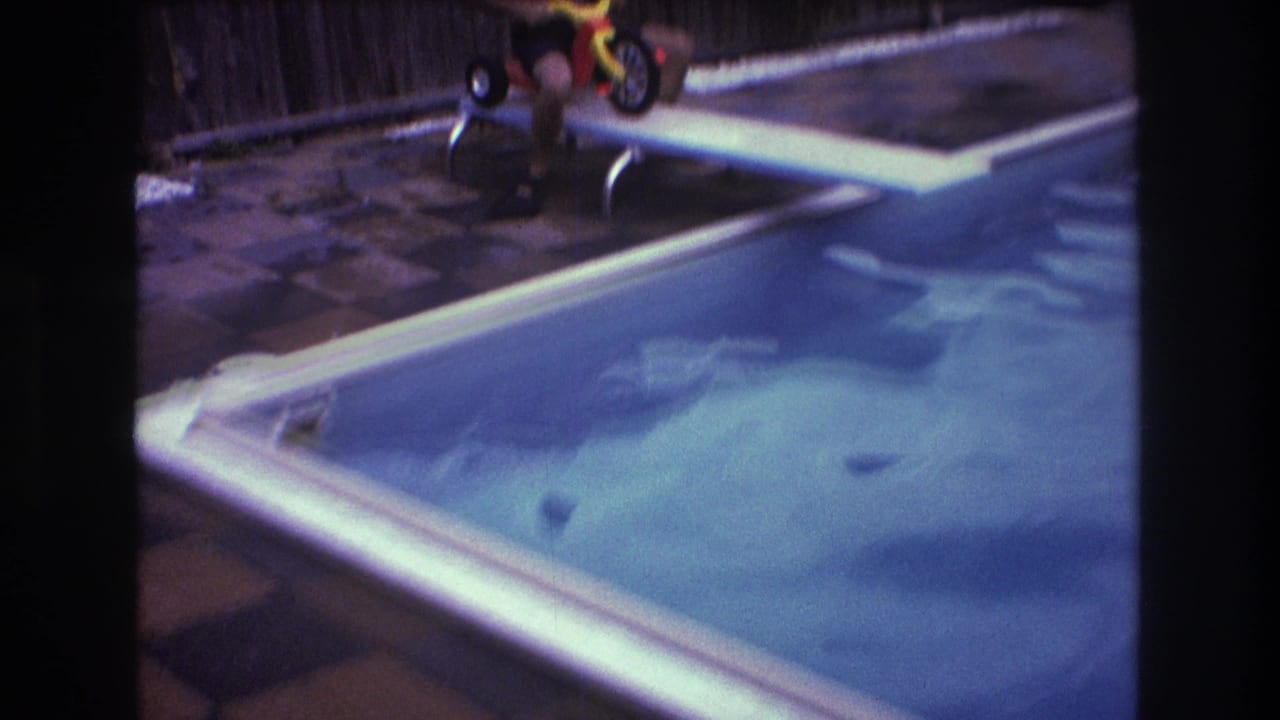 In a 1975 Boston 8mm home-movie frame, a shirtless man in goggles sits on a red and yellow tricycle near a white diving board over a backyard pool.