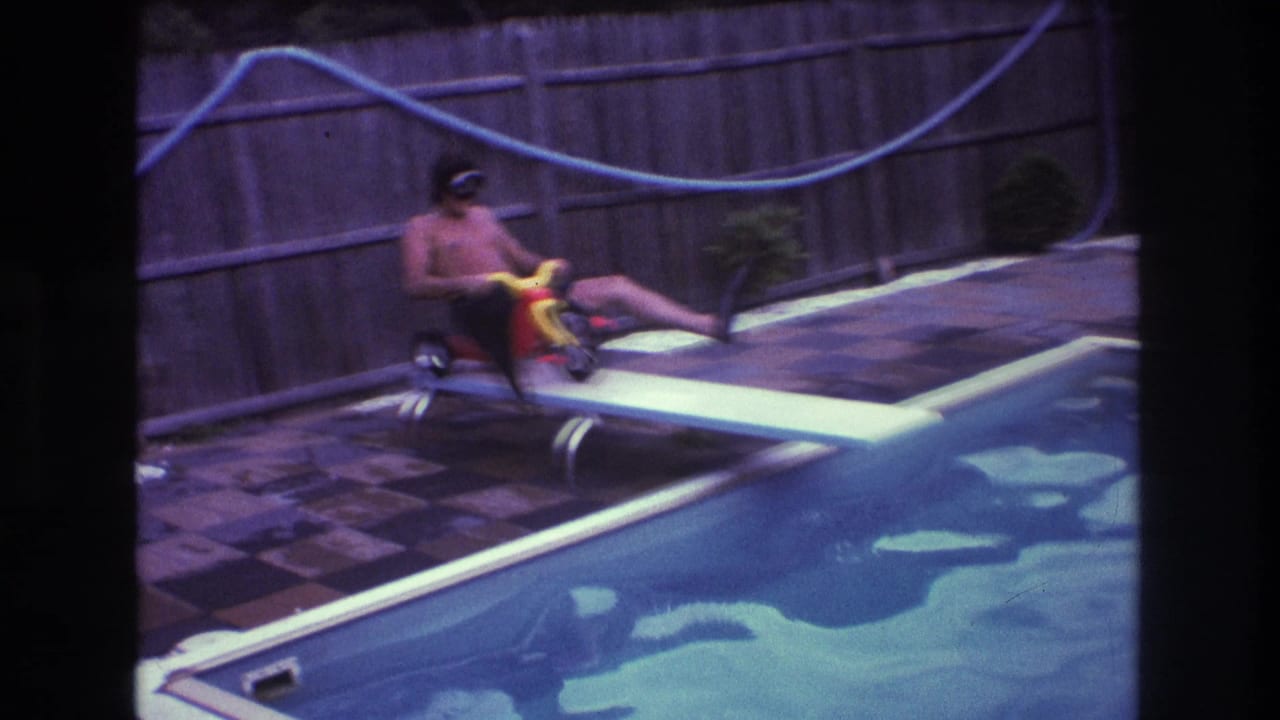 Man in goggles sits on a small tricycle on a diving board over a pool.