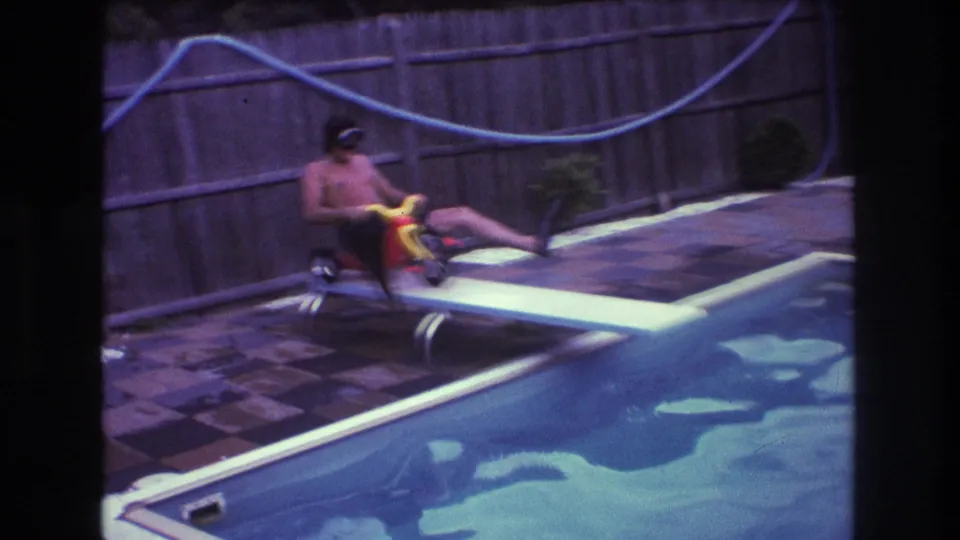 The man in goggles and swim trunks balances a small red and yellow tricycle on the diving board, with pool water, patio tiles, fence, and blue hose behind him.