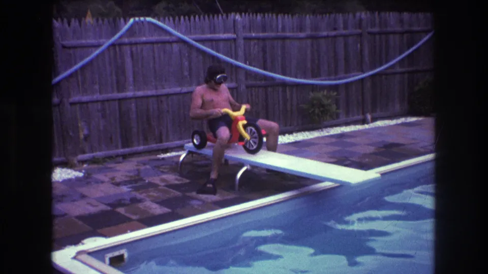 A 1970s home-movie frame shows the shirtless man holding the handlebars of the small tricycle while seated near the end of the white diving board over the pool.