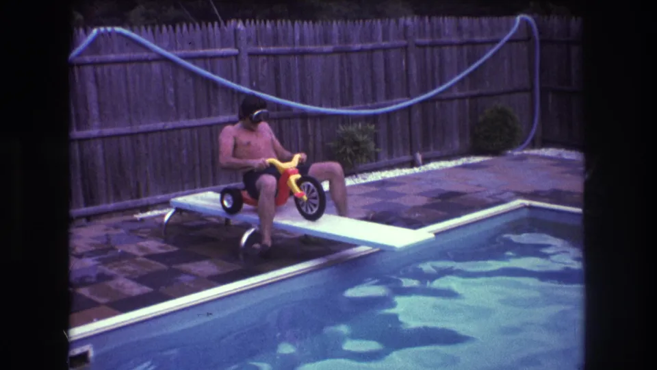 The man in swim trunks and goggles sits on the red and yellow tricycle on the diving board, framed by rippling pool water and a fenced backyard patio.