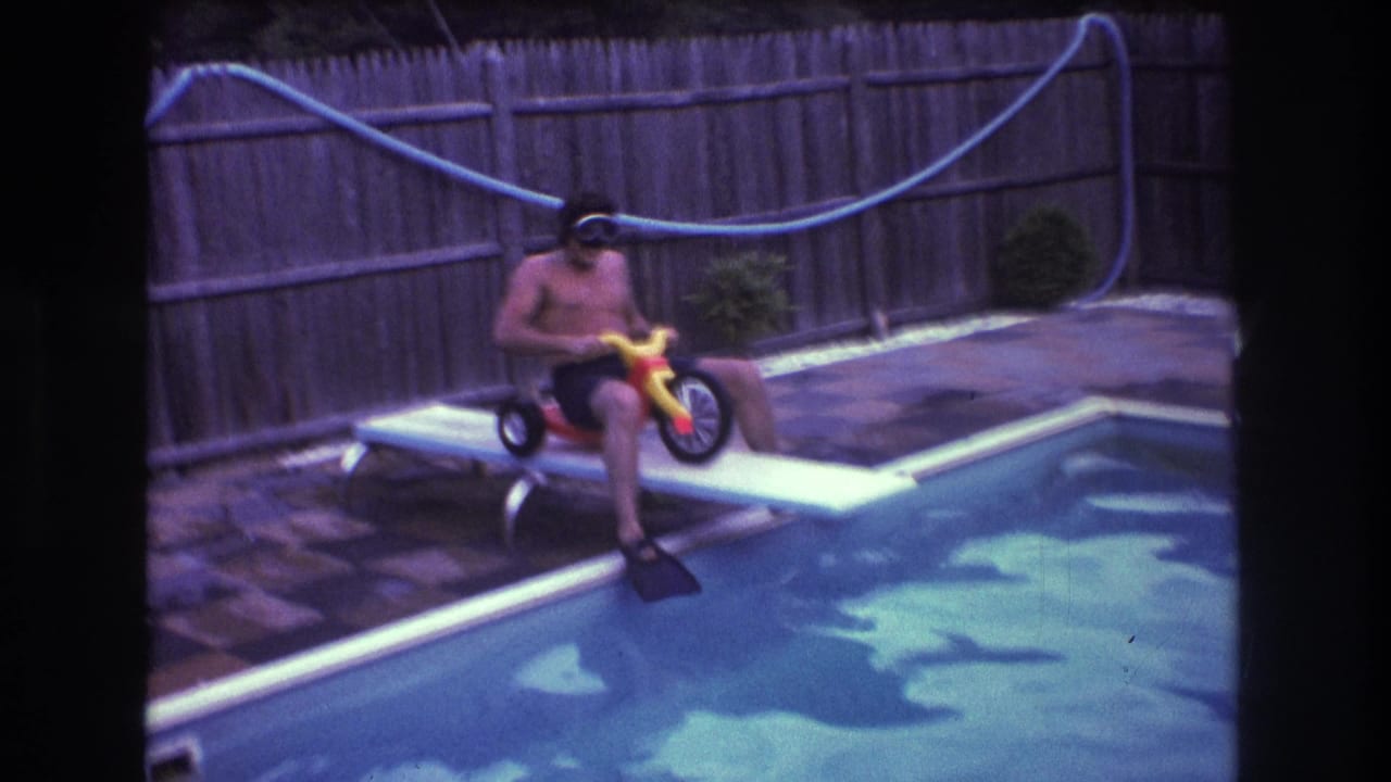 In 1975 Boston-area 8mm home-movie footage, an adult man wearing goggles, swim cap, and flippers sits on a red and yellow tricycle on a backyard pool diving board.