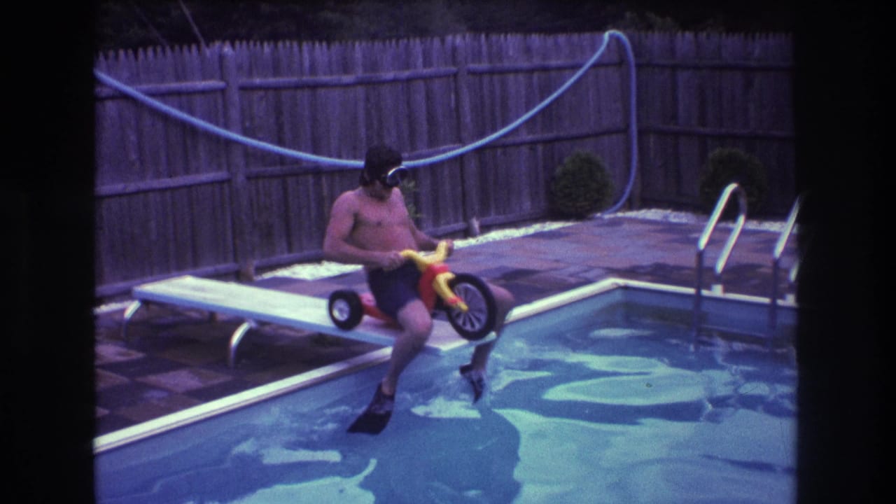 Man on a small tricycle rolls forward on the diving board beside the swimming pool.