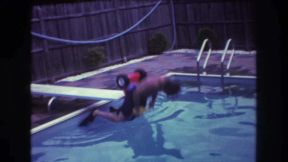 The rider falls forward from the diving board as the small tricycle angles toward the swimming pool, with the fence, hose, and pool ladder in the background.