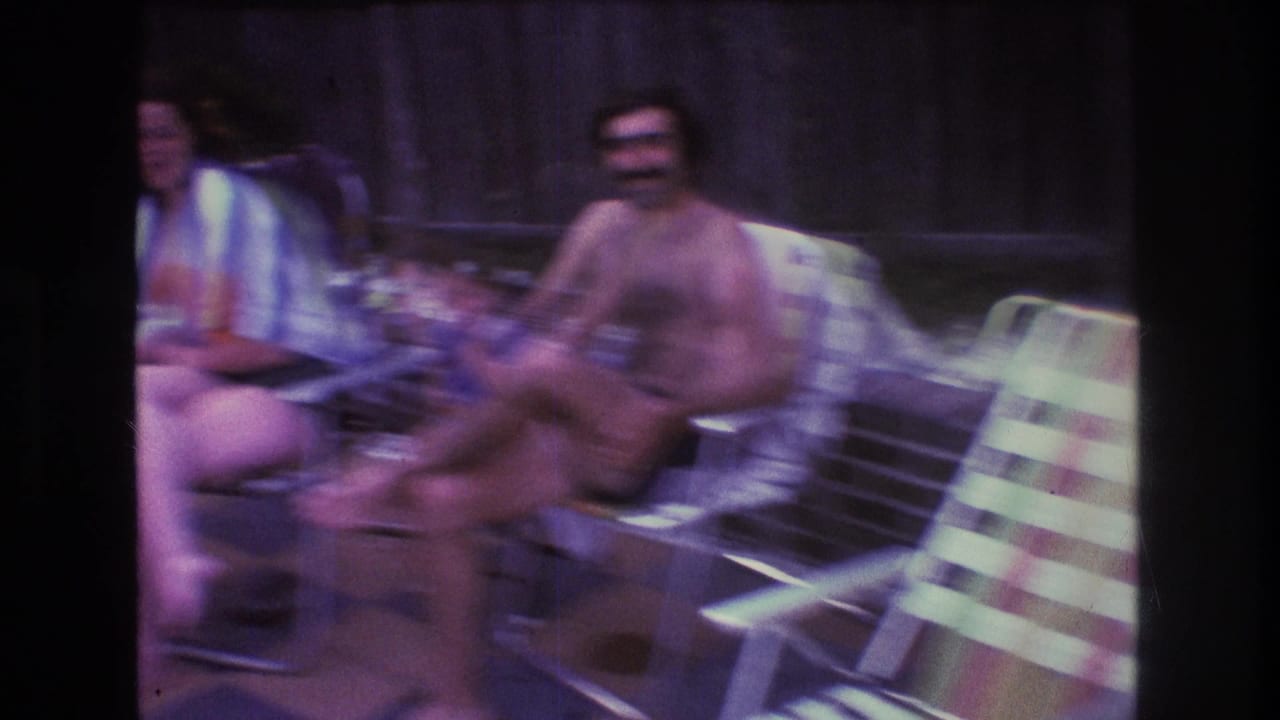 1975 Boston-area 8mm home-movie frame showing adults seated on metal patio chairs near a wooden fence during a casual outdoor gathering.