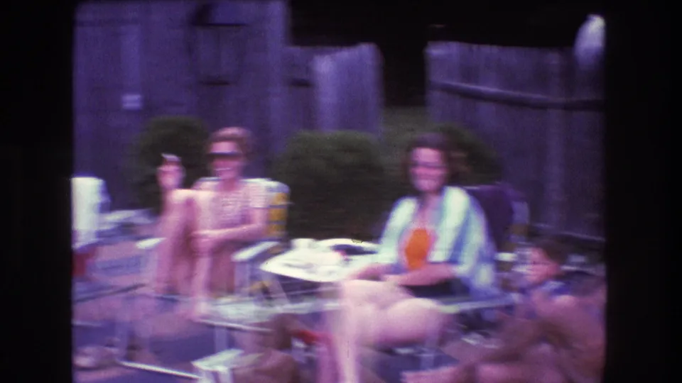 1975 Boston-area 8mm frame showing several adults seated around retro patio furniture, including a small round table and checkered patio surface.