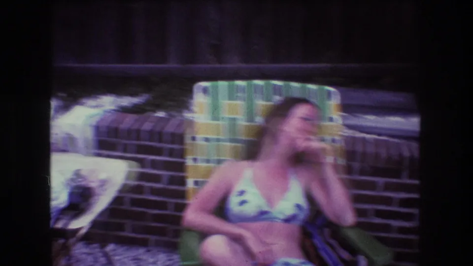 1975 Boston-area home-movie frame showing a woman in a patterned bikini reclining in a green patio chair near brick edging and a wooden fence.