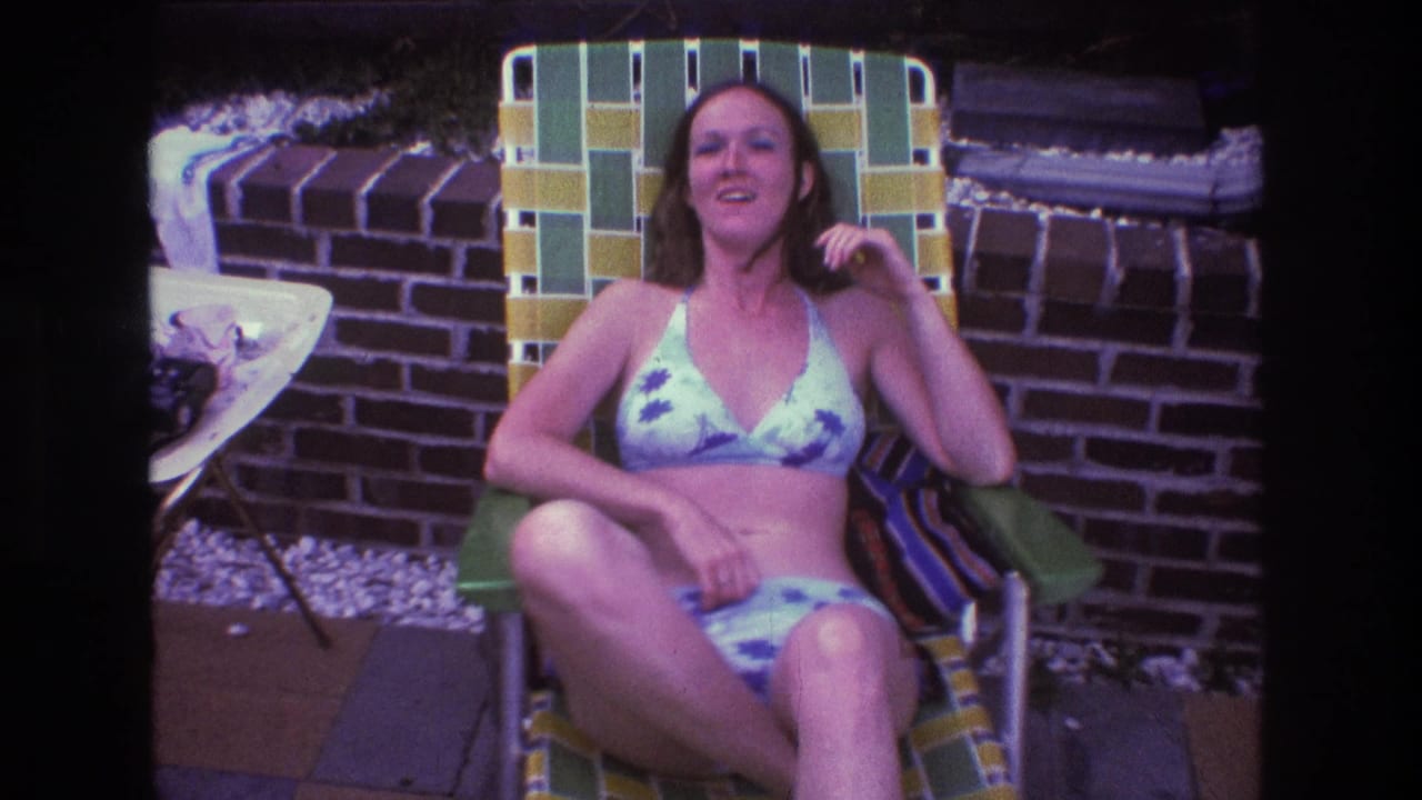 In 1975 Boston home-movie footage, a woman in a light patterned bikini smiles while seated in a green and yellow woven lawn chair beside a brick wall.