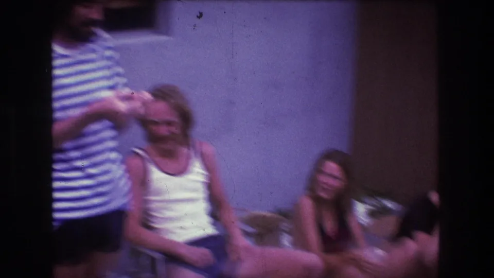 In the backyard gathering, a man in a blue and white striped shirt stands near a seated woman in a white tank top and another woman seated beside her.