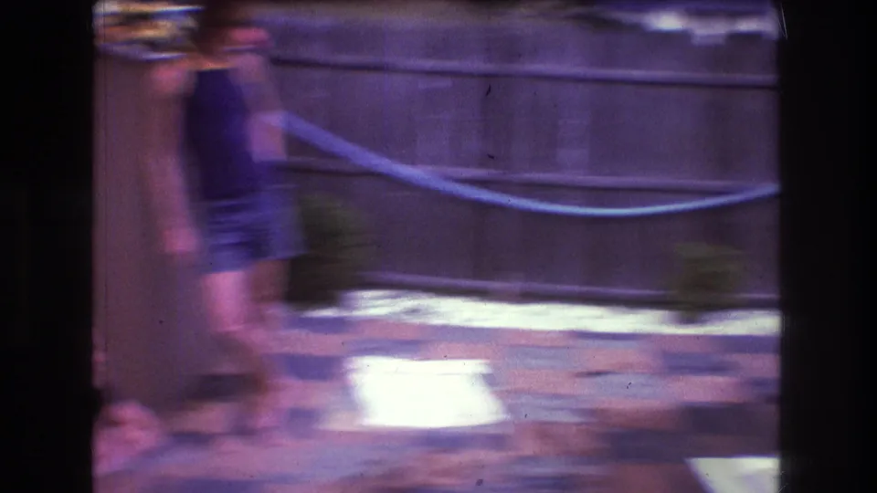 A motion-blurred figure crosses the patio in front of a wooden fence, with handheld camera movement and faded color typical of the 8mm backyard scene.