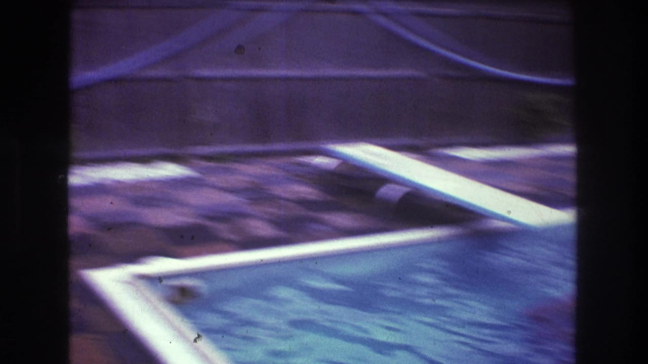 1975 Boston home-movie frame showing a backyard swimming pool, diving board, tile patio, and dark film borders with no person clearly visible in this frame.