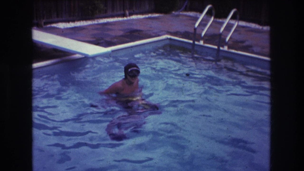 1975 Boston home-movie frame showing a swimmer in a backyard pool with a swim cap or goggles, pool ladder, tiled patio, and wooden fence visible behind.
