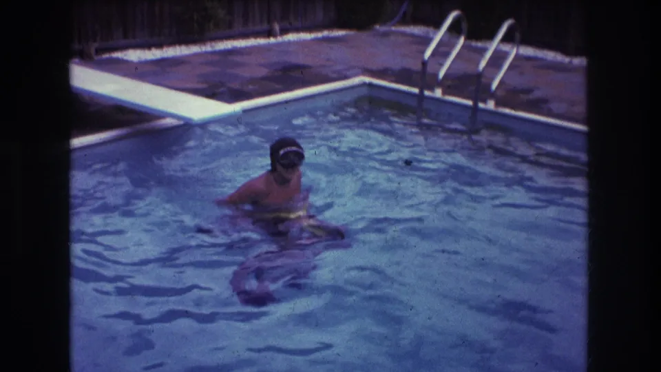 1975 Boston home-movie frame showing a swimmer in a backyard pool with a swim cap or goggles, pool ladder, tiled patio, and wooden fence visible behind.