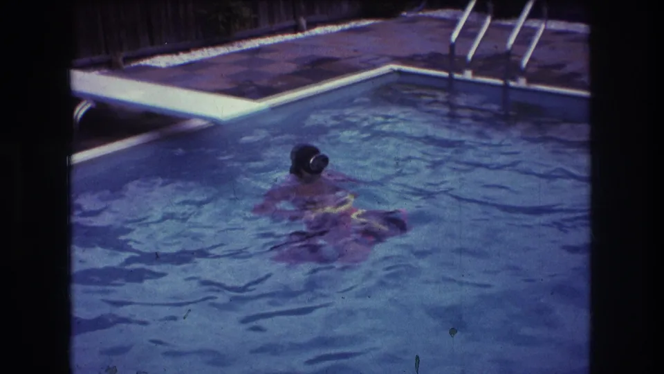 1975 Boston home-movie frame showing a swimmer floating or swimming in blue pool water, with the diving board, pool ladder, patio, and fence in view.