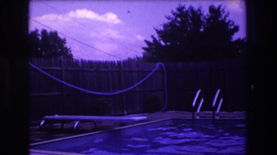 1975 Boston home-movie frame showing the backyard pool area without a visible person, including the pool ladder, wooden fence, lounge chair, hose or pool line, and trees against the sky.