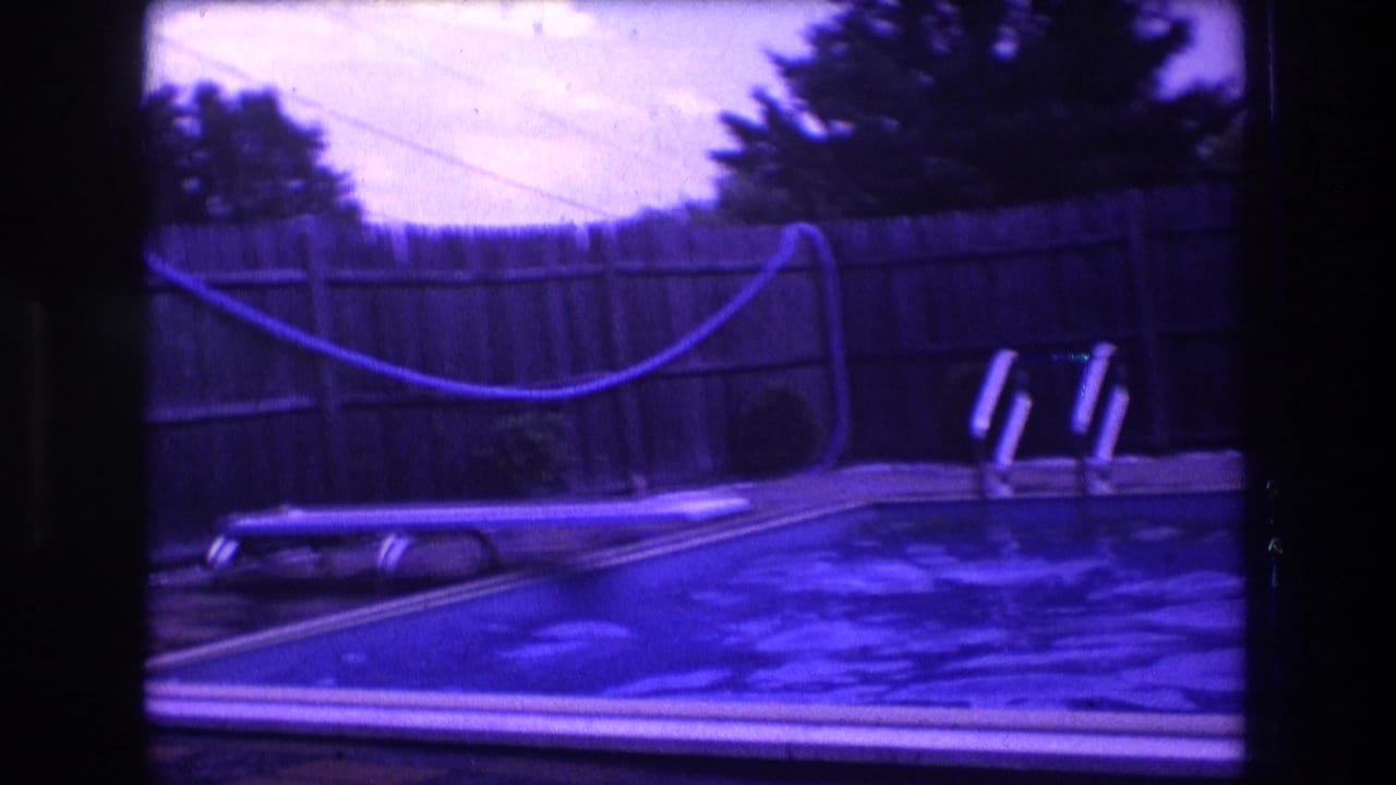 1975 Boston home-movie frame showing a backyard swimming pool, lounge chair, wooden privacy fence, trees, and a coiled garden hose under a strong purple-blue film cast.
