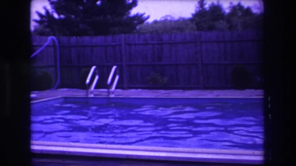 1975 Boston home-movie still of a backyard pool with metal ladder, wooden fence, hose, trees, and visible film grain and gate edge.