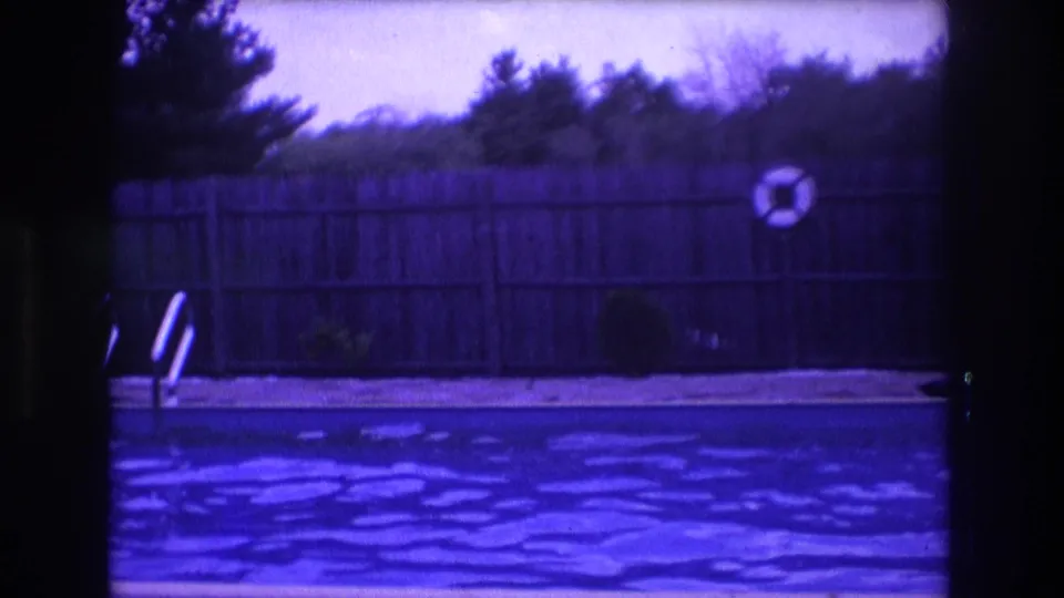 1975 Boston backyard pool frame showing rippling water, a wooden privacy fence, partial metal ladder, shrubs, trees, and a small flotation ring near the fence.