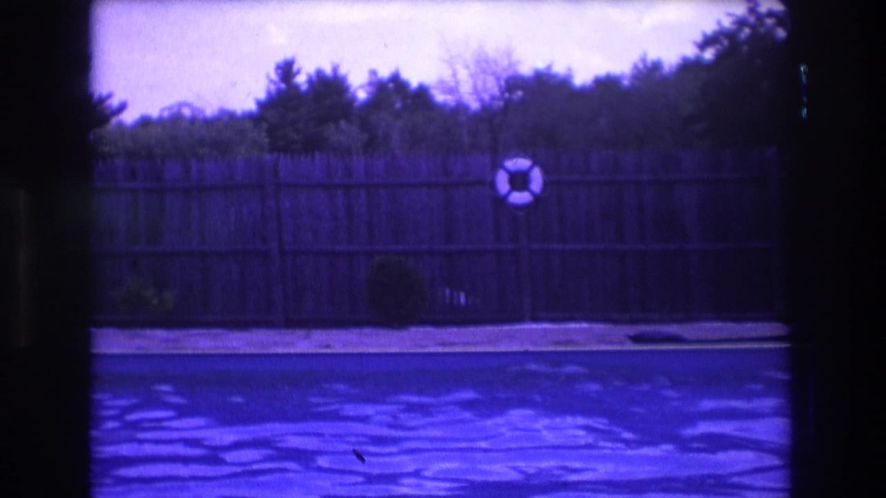1975 Boston 8mm frame showing rippling backyard pool water, a wooden fence, a mounted white life preserver, shrubs, and dark film-edge borders.