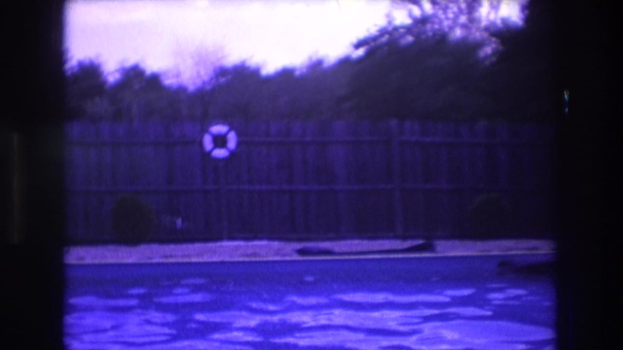 1975 Boston home-movie frame with blue-purple pool water, wooden fence, centered life preserver, shrubs, and a dark object along the pool edge.