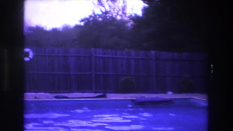 1975 Boston 8mm frame during a pan across a backyard pool, with rippling water, a long wooden fence, shrubs, and a partially visible life preserver at left.