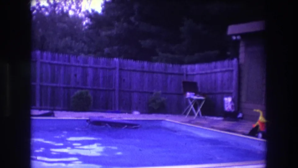 1975 Boston home-movie frame showing the pool corner, wooden fence, shrubs, a small poolside table or chair, a shed edge, and a yellow toy near the right side.