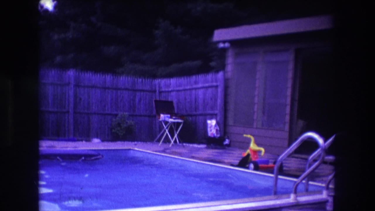 1975 Boston home-movie frame showing a blue inground pool, wood privacy fence, small shed, patio area, metal ladder, and a poolside toy.
