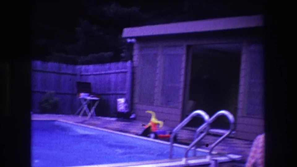 1975 Boston backyard poolside frame with metal ladder rails in the foreground, a wood fence, small shed, folding chair, and toy near the pool edge.