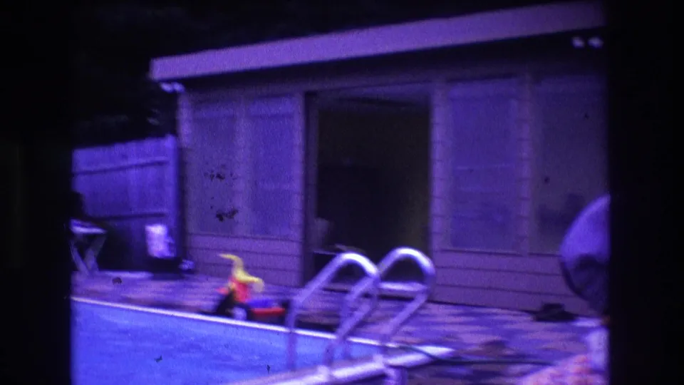 1975 Boston home-movie frame moving closer to the pool ladder and shed, with the wood fence, patio, pool water, and scattered poolside items visible.