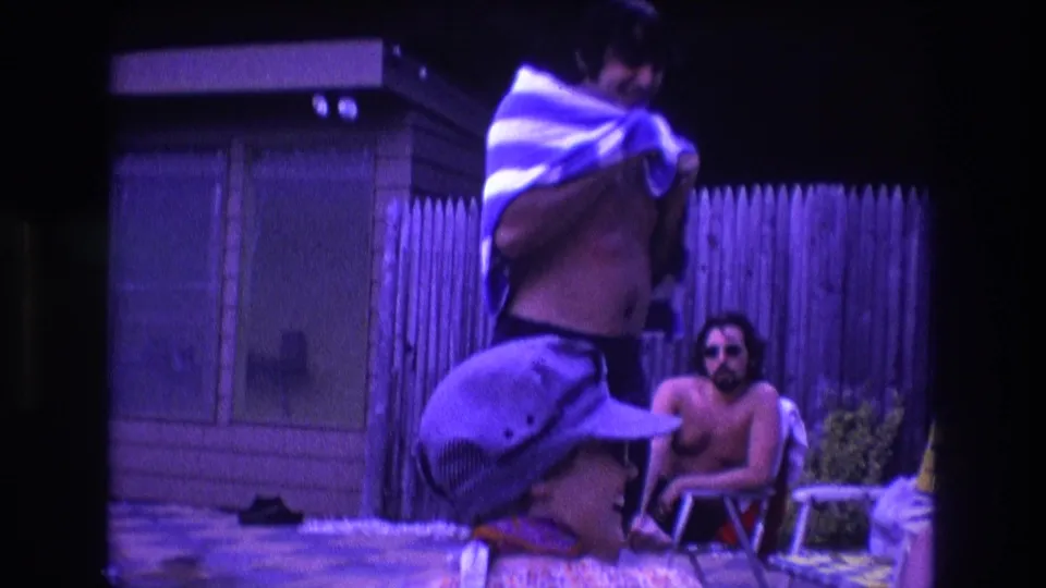 1975 Boston poolside home-movie frame showing an adult man standing with a striped towel and another adult man seated in sunglasses near the fence and shed.